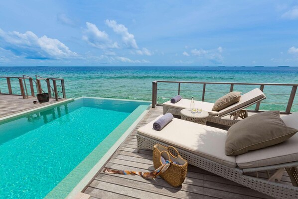 Zona junto a la piscina con sillones de descanso y una terraza de madera con vistas al mar azul y cristalino.