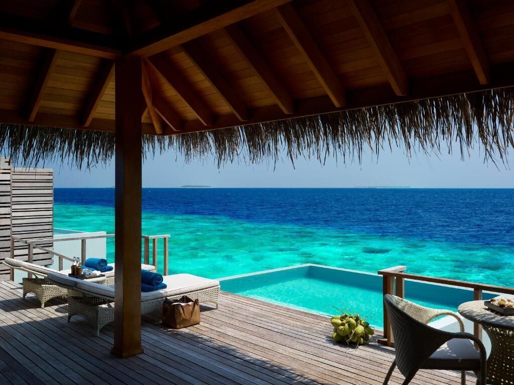 Terraza de madera con sillones de descanso, una pequeña mesa y una piscina con vistas al mar azul cristalino.