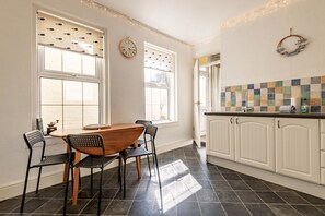 A kitchen with white cabinets, a tiled backsplash, and a dining area with a wooden table and chairs. - Kirkmay Road (No 18)  Mayfield House | Crail (Crail)