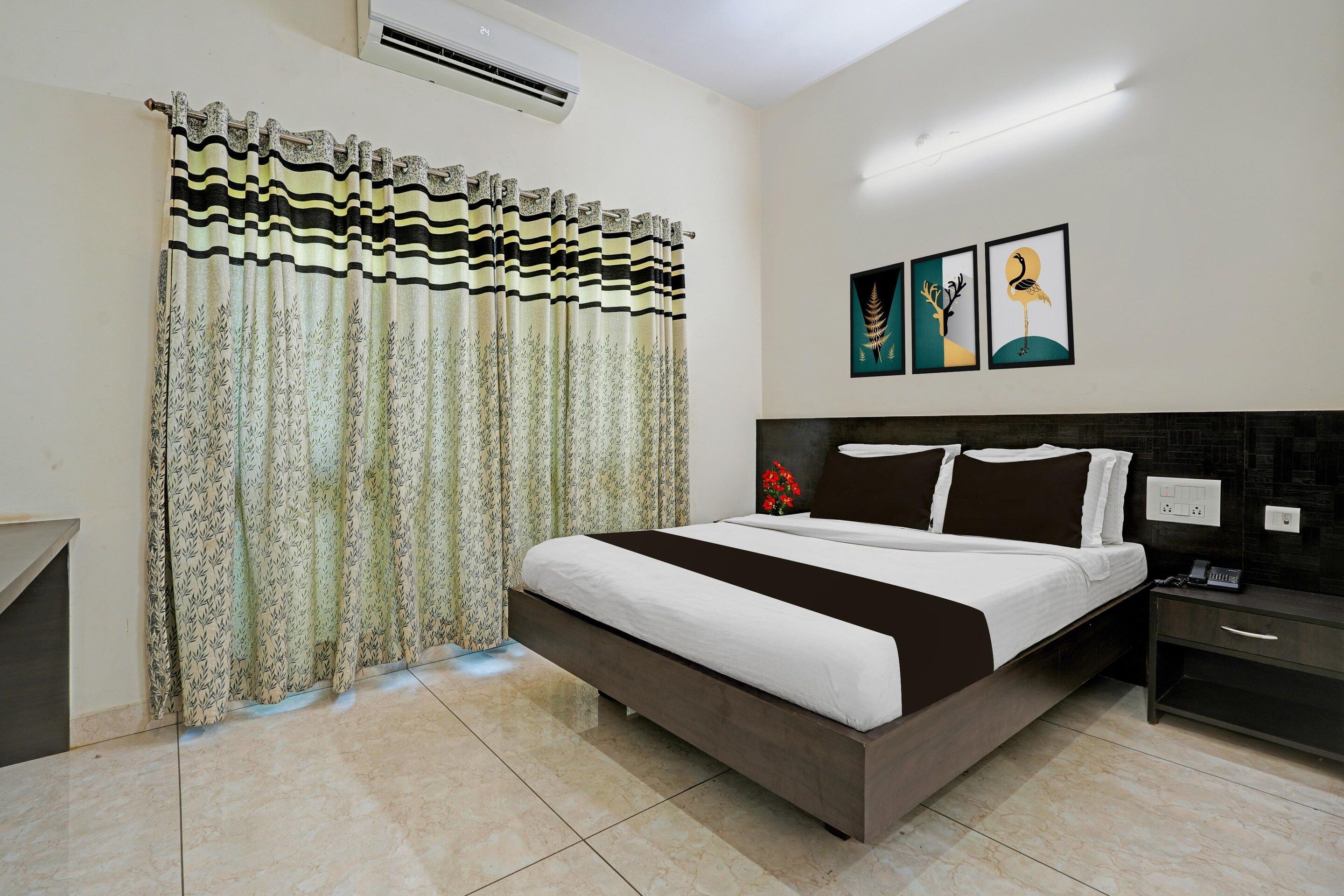 A hotel room with a bed, a nightstand, a wall-mounted air conditioner, and a window with patterned curtains.