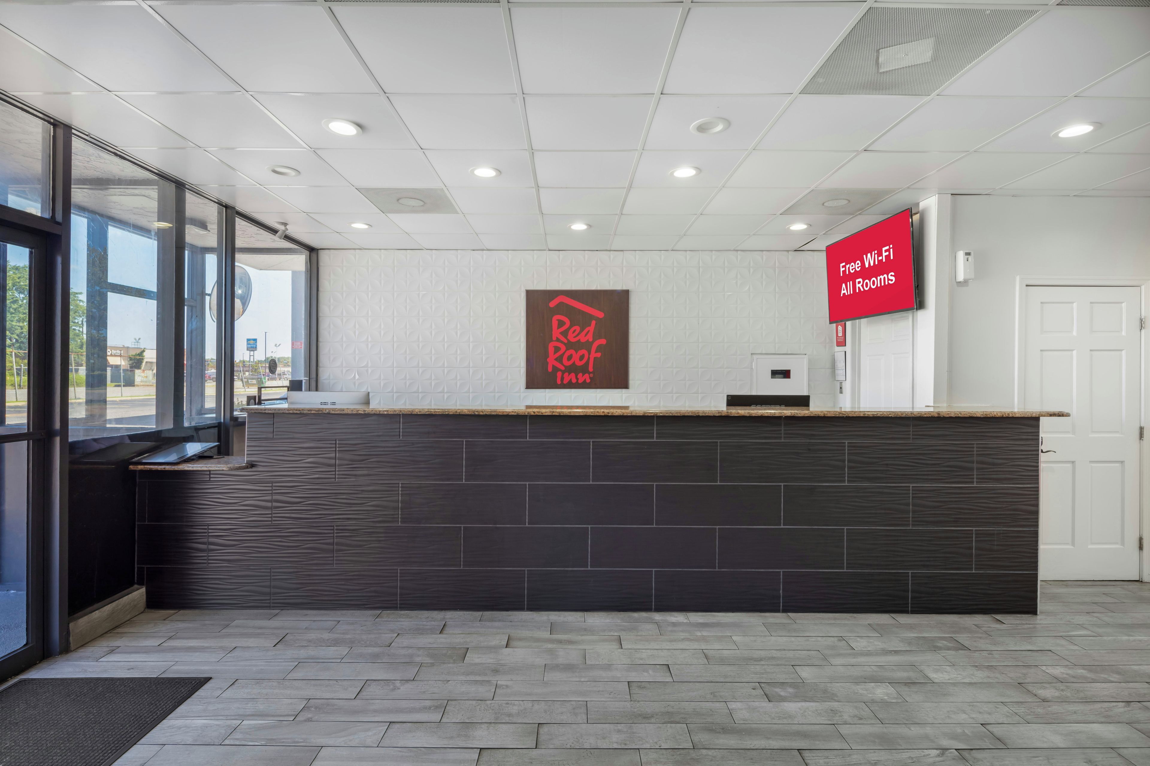 A reception desk with a Red Roof Inn sign, a digital display advertising free Wi-Fi, and a view of the outside through large windows.