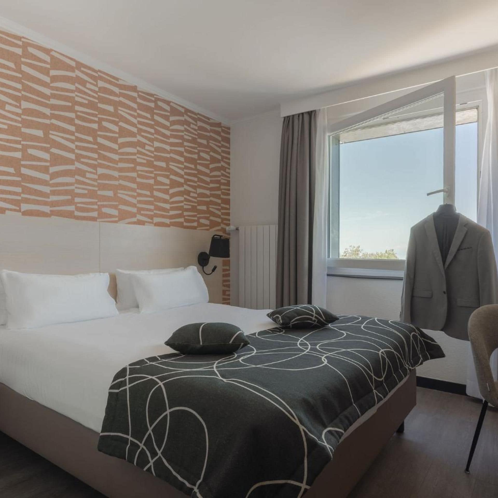 A hotel room with a bed, a window with a view, a coat rack, and a wall with a patterned design.