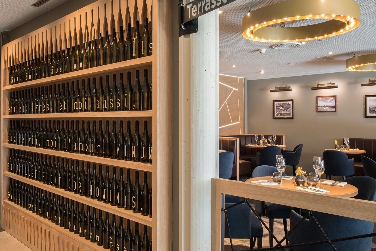 A restaurant interior with a wall of wine bottles, tables with chairs, and a sign reading 'Terrasse'.
