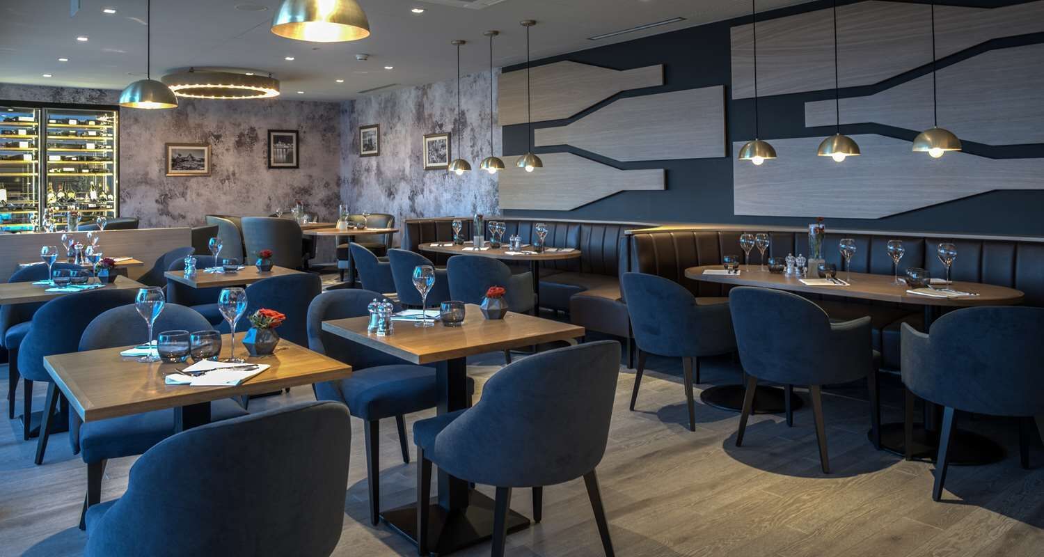 A modern restaurant interior with multiple dining tables and chairs, pendant lighting, and a bar area with shelves.