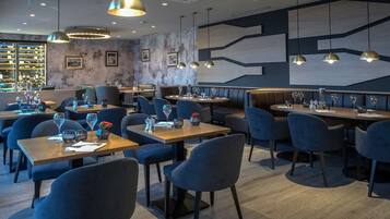 A modern restaurant interior with multiple dining tables and chairs, pendant lighting, and a bar area with shelves.