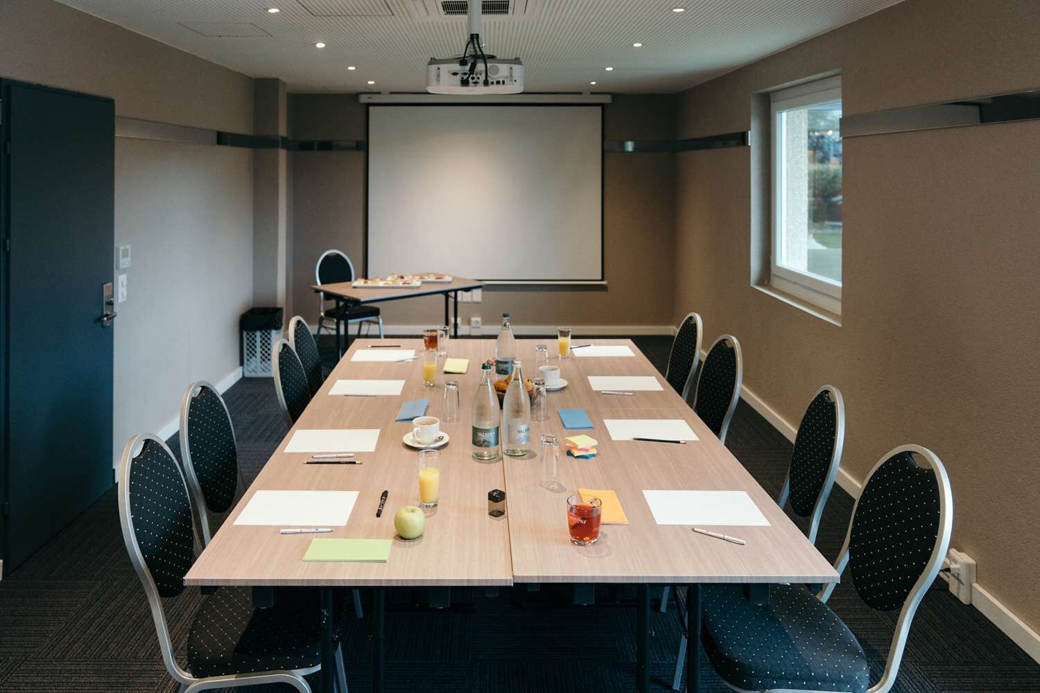 Ein Konferenzraum mit langem Tisch, Stühlen, einer Projektionsleinwand und verschiedenen Gegenständen auf dem Tisch.