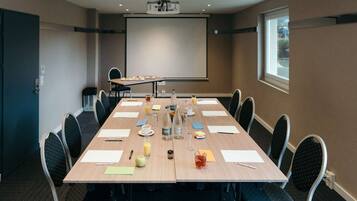 Ein Konferenzraum mit langem Tisch, Stühlen, einer Projektionsleinwand und verschiedenen Gegenständen auf dem Tisch.