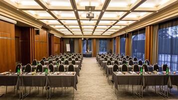 Une salle de conférence avec des rangées de tables et de chaises, un projecteur et une grande fenêtre avec des rideaux.