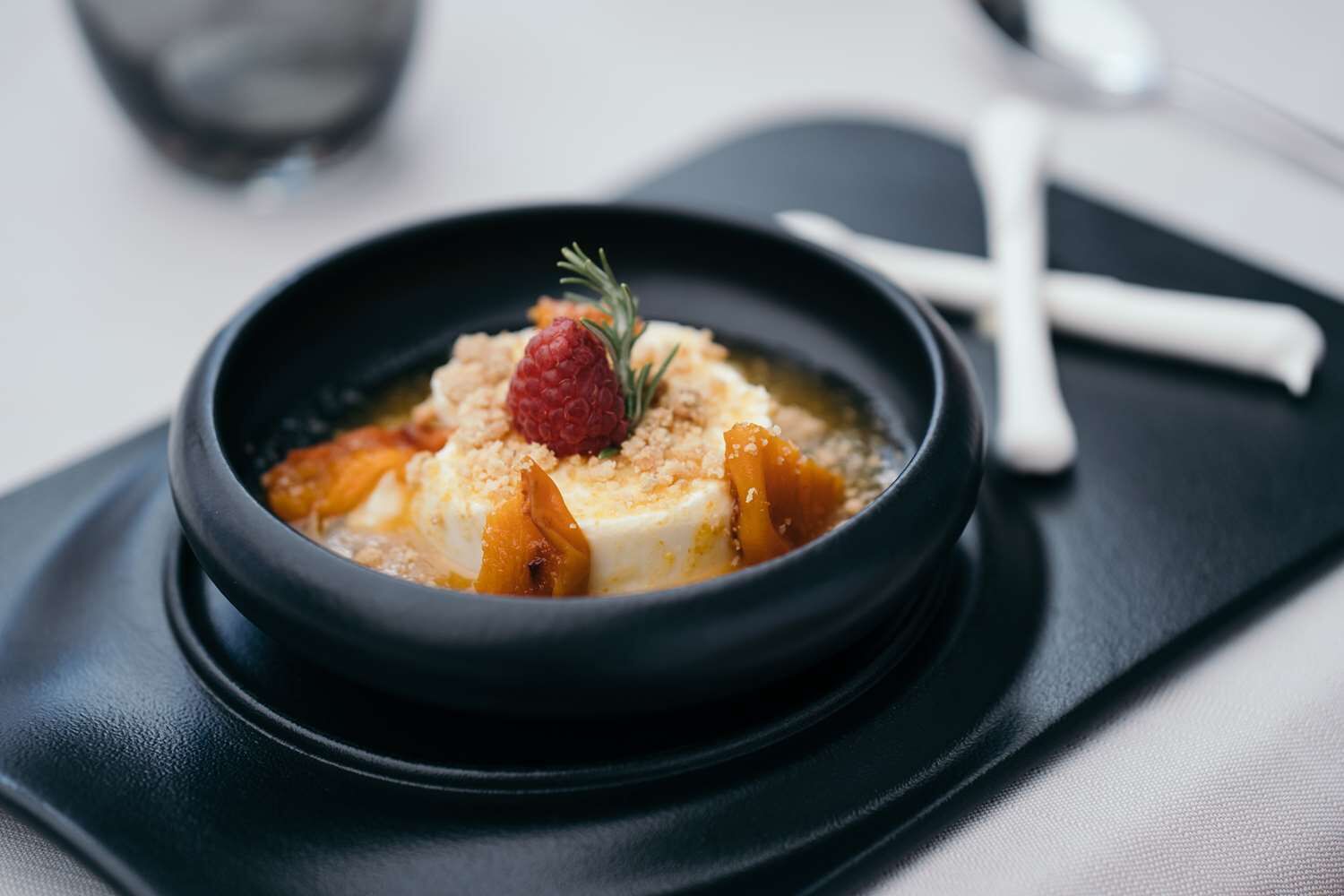 A dessert in a black bowl on a black tray with a spoon and fork beside it.
