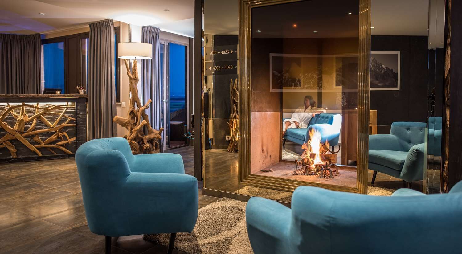 Un área de descanso moderna con sillones azules, una escultura de madera y un panel de vidrio que refleja a una persona sentada junto a una chimenea.