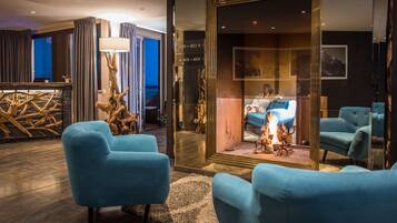 Un área de descanso moderna con sillones azules, una escultura de madera y un panel de vidrio que refleja a una persona sentada junto a una chimenea.
