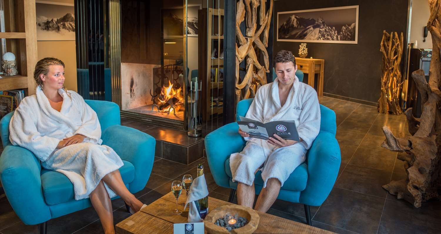 Two individuals in white bathrobes seated on blue chairs in a room with a fireplace, wooden decor, and a small table with a bottle and glasses.