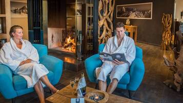 Two individuals in white bathrobes seated on blue chairs in a room with a fireplace, wooden decor, and a small table with a bottle and glasses.