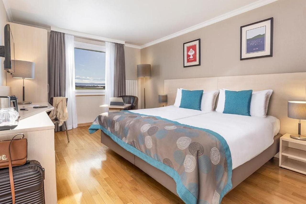 A hotel room with a bed, bedside tables, a desk, a chair, a window with curtains, and artwork on the walls.
