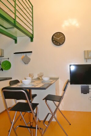 A dining area with a table set for two, a green metal staircase, a wall-mounted clock, and a flat-screen TV. - Vrbo Property (Milano)