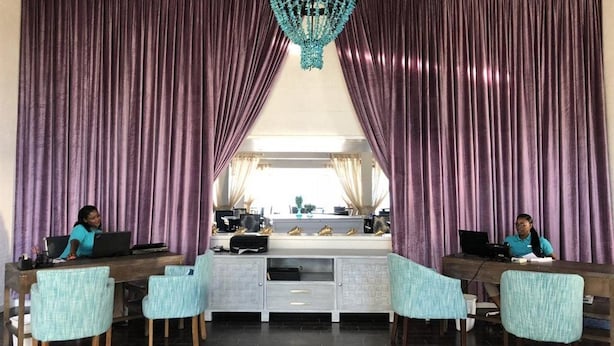 Una habitación con dos estaciones de trabajo, cada una con una computadora portátil, un escritorio y una silla. Cuenta con un ventanal con cortinas, un candelabro y un mostrador con varios objetos.