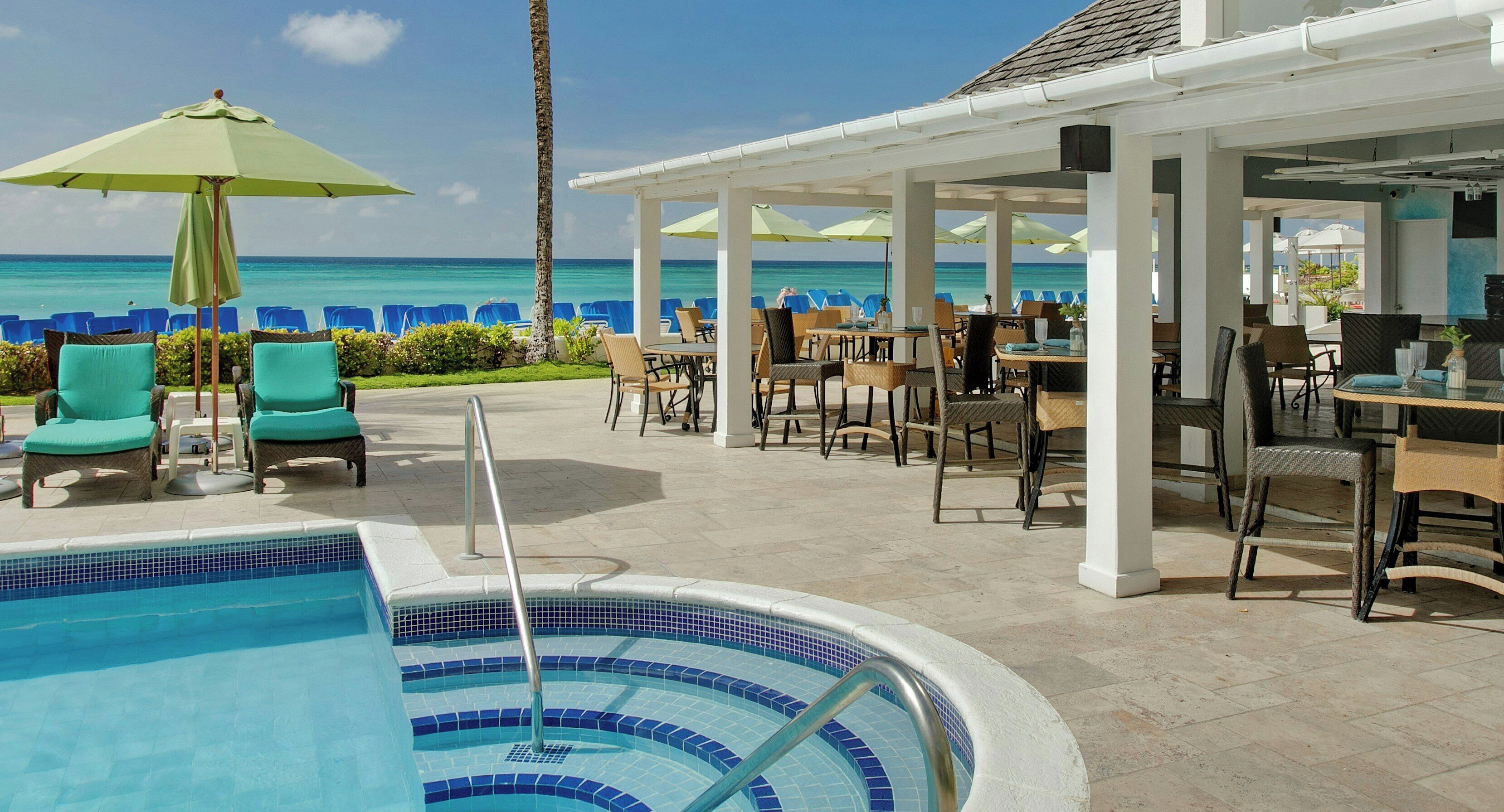 Un espace aménagé au bord de la piscine, avec des chaises longues et des parasols, une piscine aux carreaux bleus et une vue sur l’océan.