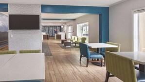 Un hall d’hôtel moderne avec un plancher en bois, un grand téléviseur à écran plat fixé sur un mur texturé, et un espace salon avec des tables et des chaises.
