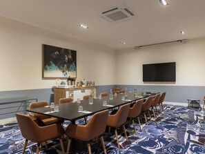 Un salón de conferencias con una larga mesa, sillas, una pantalla de proyector y un cuadro en la pared.