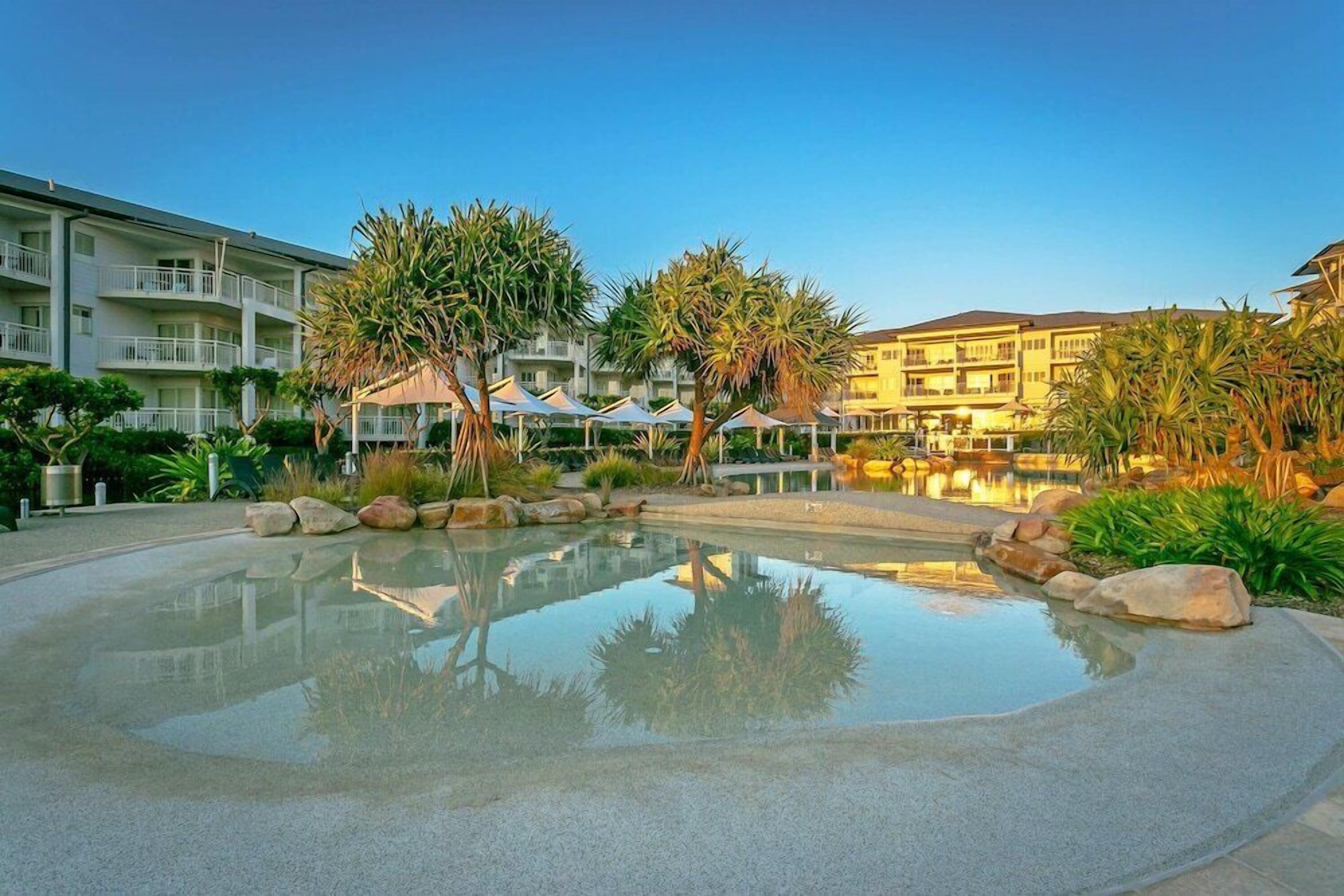 A hotel with a swimming pool, palm trees, and a clear sky.