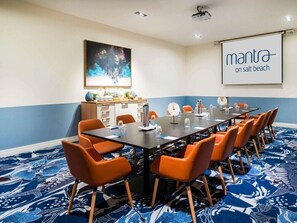 Un salón de conferencias con una larga mesa, sillas naranjas, una pantalla de proyector y un cuadro abstracto grande en la pared.