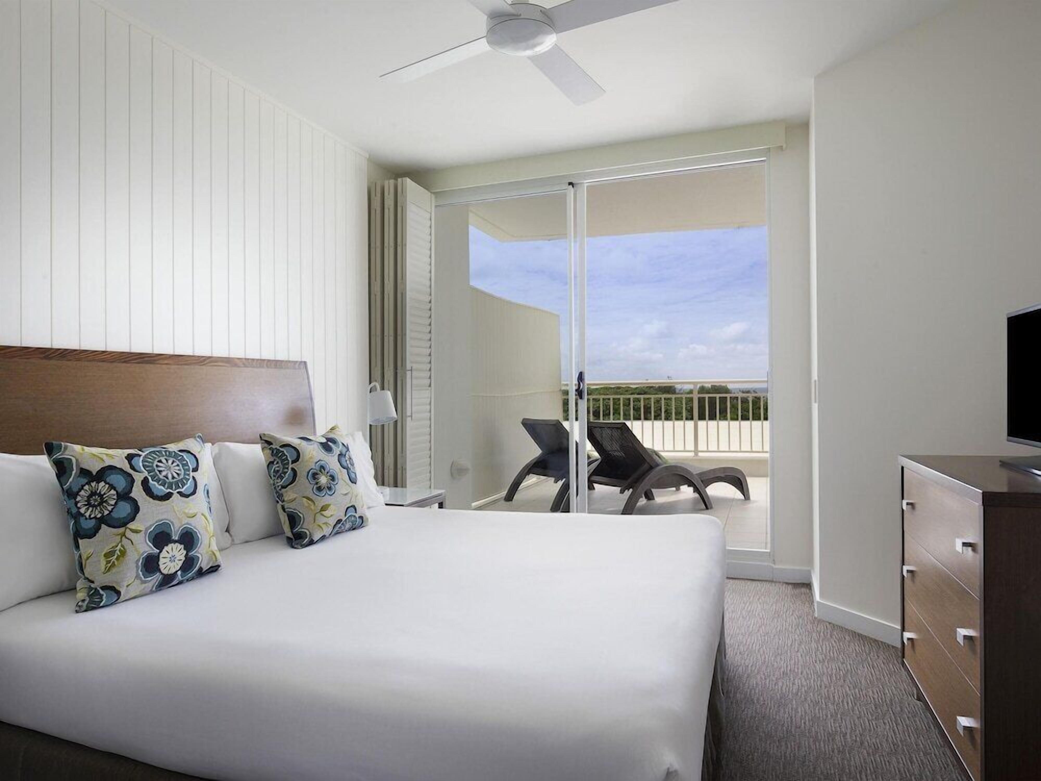 A hotel room with a bed, a dresser, a television, a ceiling fan, and a balcony with chairs.