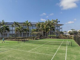 Cancha de tenis con red, rodeada de palmeras y un edificio con varios balcones.
