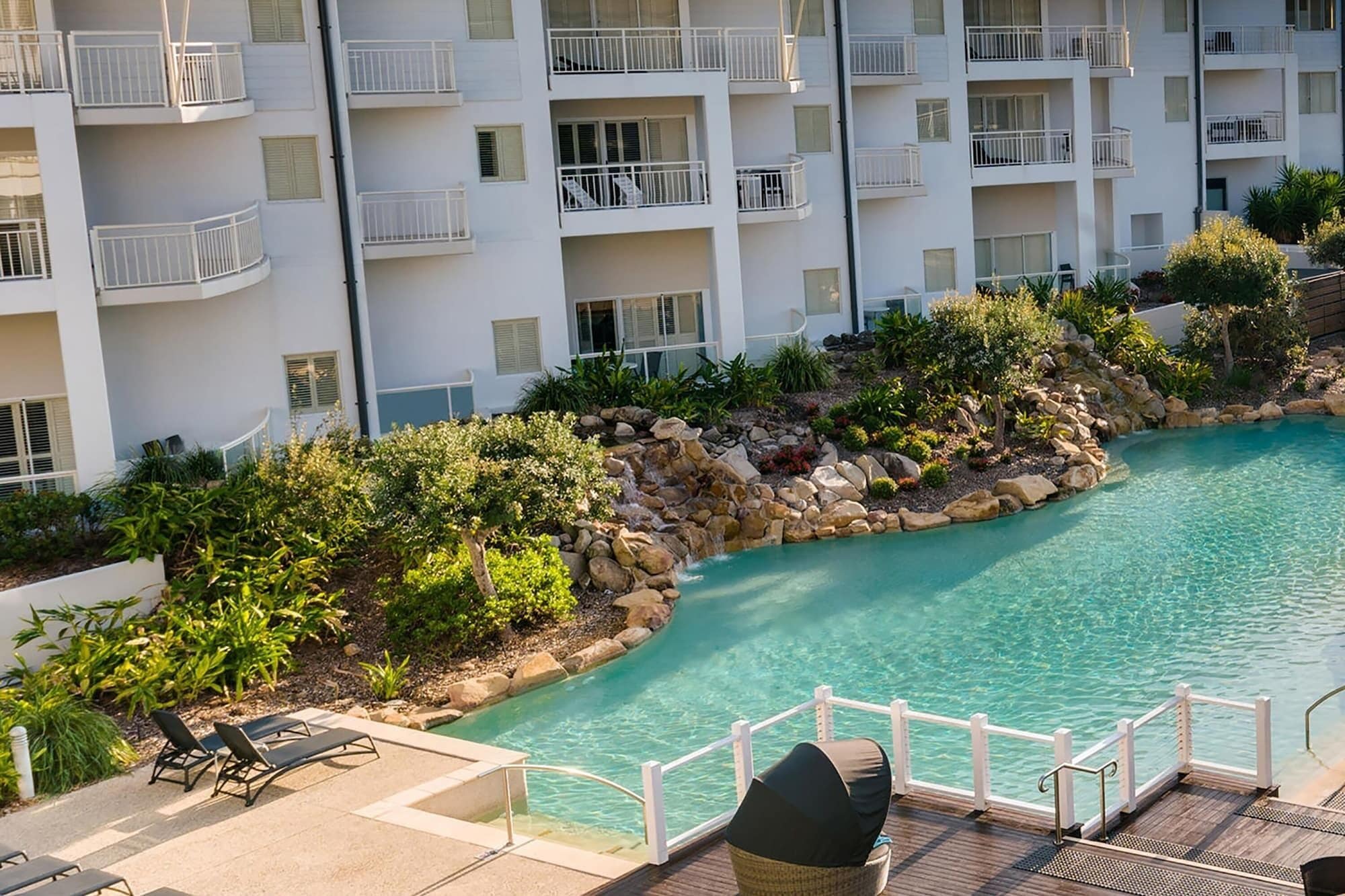 A pool surrounded by a landscaped area with a rocky edge, a wooden deck, and lounge chairs.