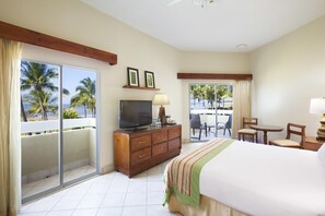 Quarto de hotel com cama, televisão, secretária e varanda vista para palmeiras.
