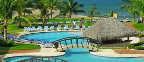 A resort pool with a thatched-roof structure, palm trees, and lounge chairs.