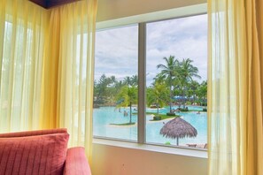 A room with a large window overlooking a pool area with palm trees and a thatched umbrella.