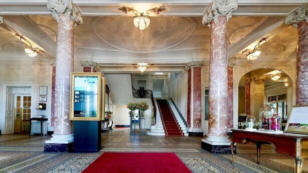 Un'imponente hall con colonne in marmo, un tappeto rosso e una scalinata che conduce ai piani superiori.