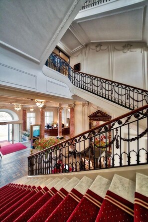 A grand staircase with ornate railings leading to a second floor, a reception area with a television, and a red carpeted area. - Schweizerhof (Lucerne)