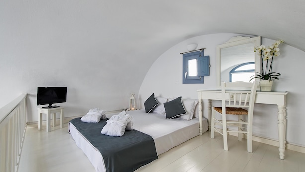 A bedroom with a bed, a desk, a chair, a TV, and a mirror. - Apanemo Hotel and Suites (Santorini)