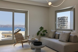 A modern living room with a sofa, a chair, a round table, and a view of the sea through large windows. - Apanemo Hotel and Suites (Santorini)