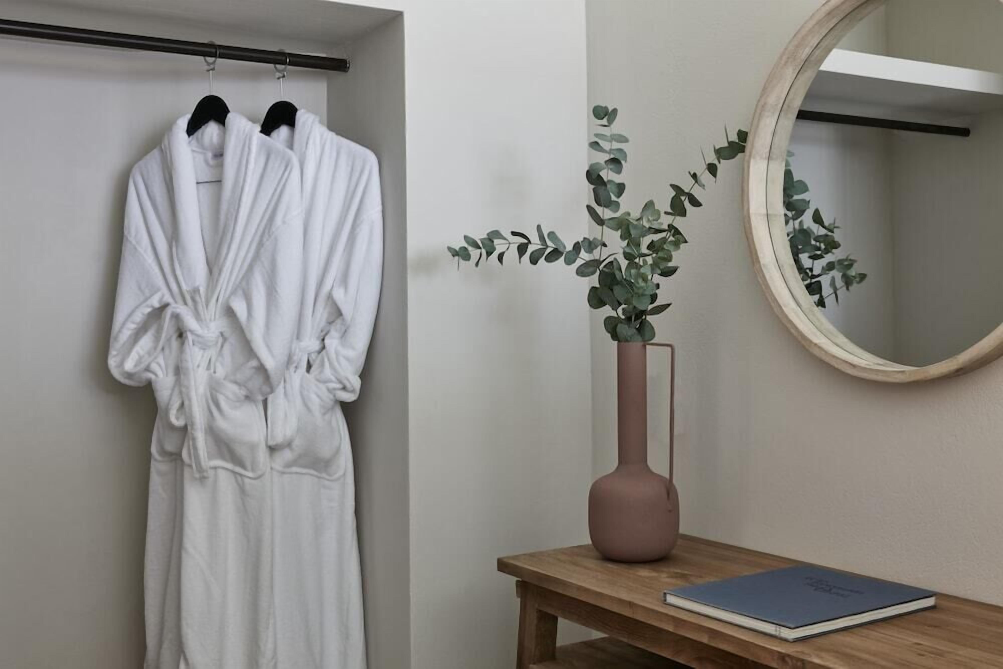 A bathroom with a towel rack holding two white bathrobes, a wooden bench with a vase of green foliage, and a round mirror on the wall.