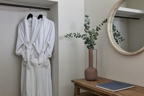 A bathroom with a towel rack holding two white bathrobes, a wooden bench with a vase of green foliage, and a round mirror on the wall. - Apanemo Hotel and Suites (Santorini)