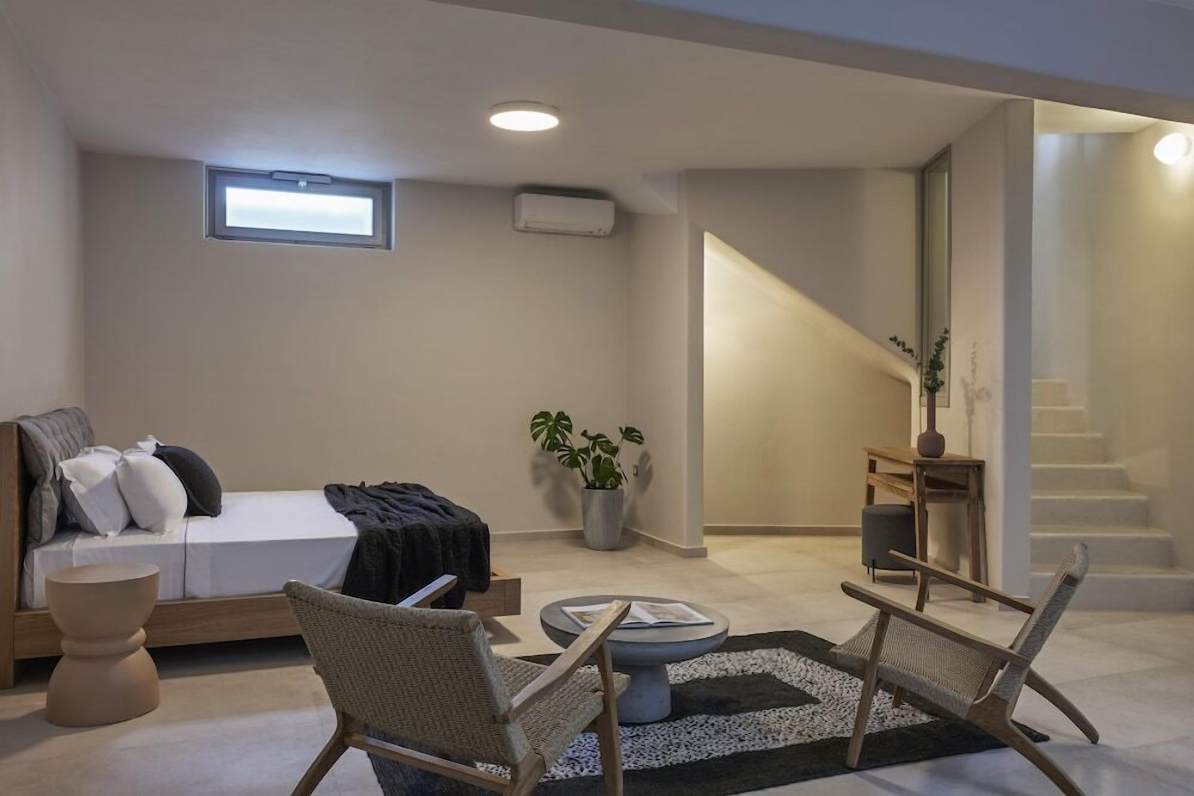 A modern bedroom with a bed, two chairs, a small table, a potted plant, and a staircase.
