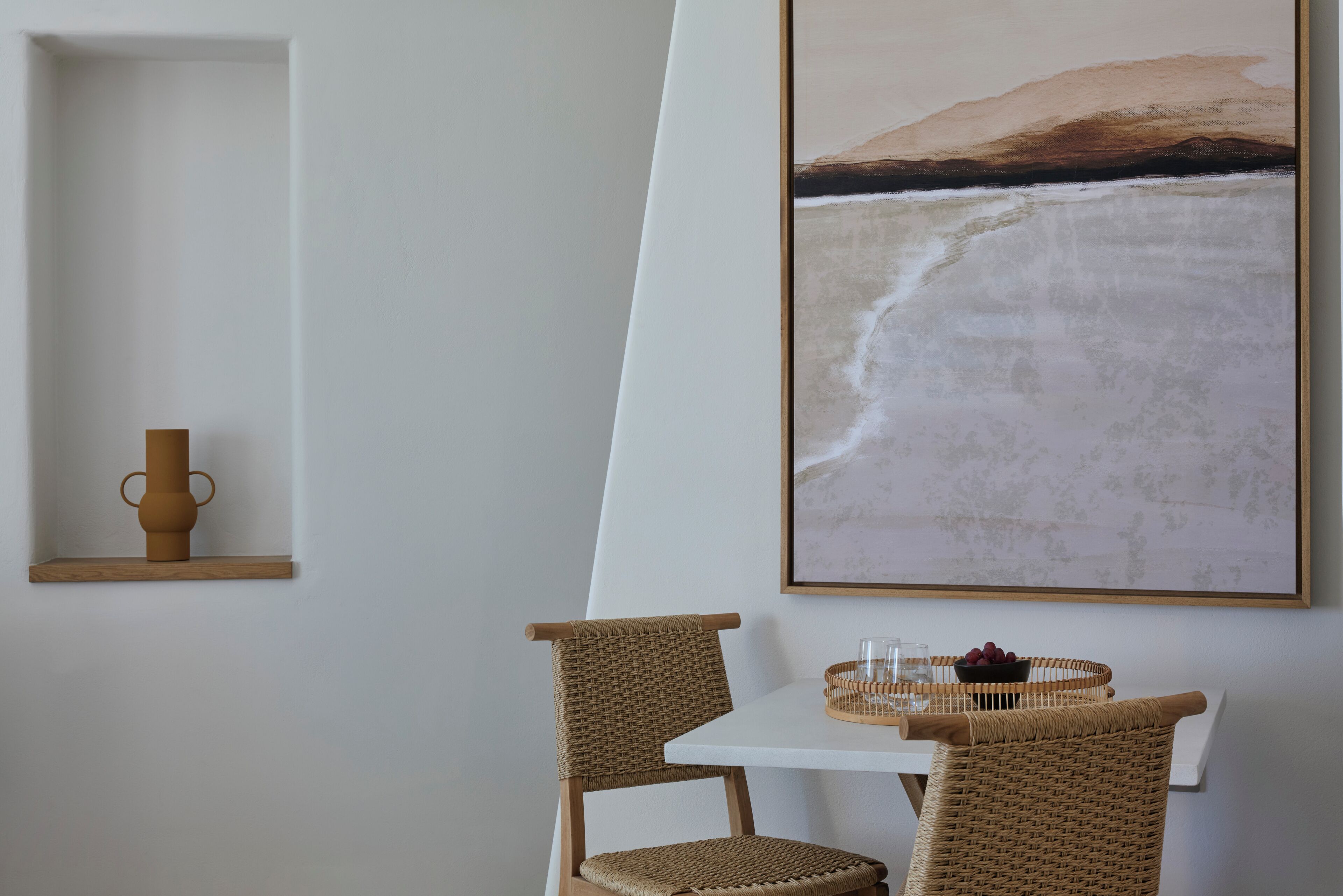 Un comedor con una mesa blanca, dos sillas de mimbre, un florero y un cuadro enmarcado de una playa.