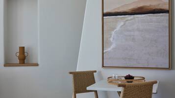 Un comedor con una mesa blanca, dos sillas de mimbre, un florero y un cuadro enmarcado de una playa.