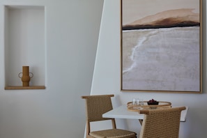 A dining area with a white table, two wicker chairs, a vase, and a framed painting of a beach scene.
