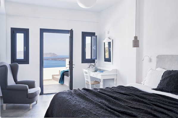A bedroom with a bed, a desk, and a chair. There are two windows with a view of the sea.