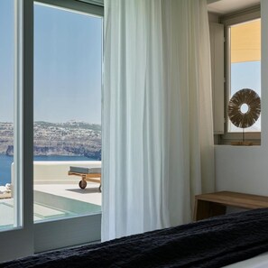 A room with a large window, a bed, a bedside table, and a view of a coastline. - Apanemo Hotel and Suites (Santorini)