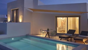 A modern outdoor pool area with a dining table and chairs, a lounge area with sun loungers, and a building entrance with glass doors. - Apanemo Hotel and Suites (Santorini)