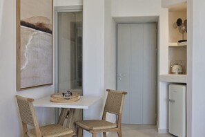 A small room with a white table, two wicker chairs, a door, and a white refrigerator. - Apanemo Hotel and Suites (Santorini)
