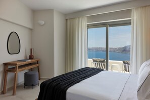 Une chambre d’hôtel avec un grand lit, un bureau avec une chaise, un petit tabouret et une vue sur la mer par une grande fenêtre.