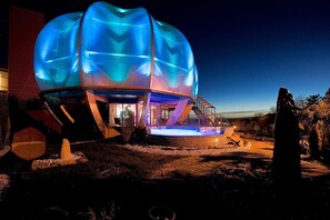 Una estructura esférica con cúpula transparente, iluminada desde el interior, y una escalera que conduce a una entrada.
