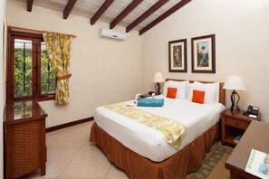 A hotel room with a bed, two bedside tables, a desk, a window with curtains, an air conditioner, and two framed pictures on the wall.