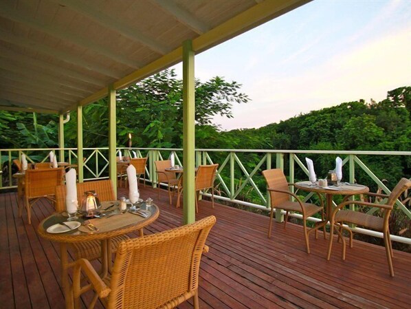 An outdoor dining area with wicker furniture and tables, surrounded by a wooden deck and a green railing.