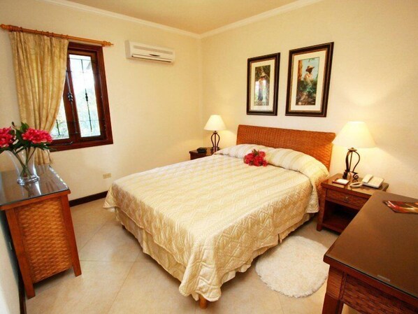 A hotel room with a bed, bedside tables, a desk, a chair, a window with curtains, and wall-mounted air conditioning. - Sugar Cane Club Hotel & Spa (Speightstown)
